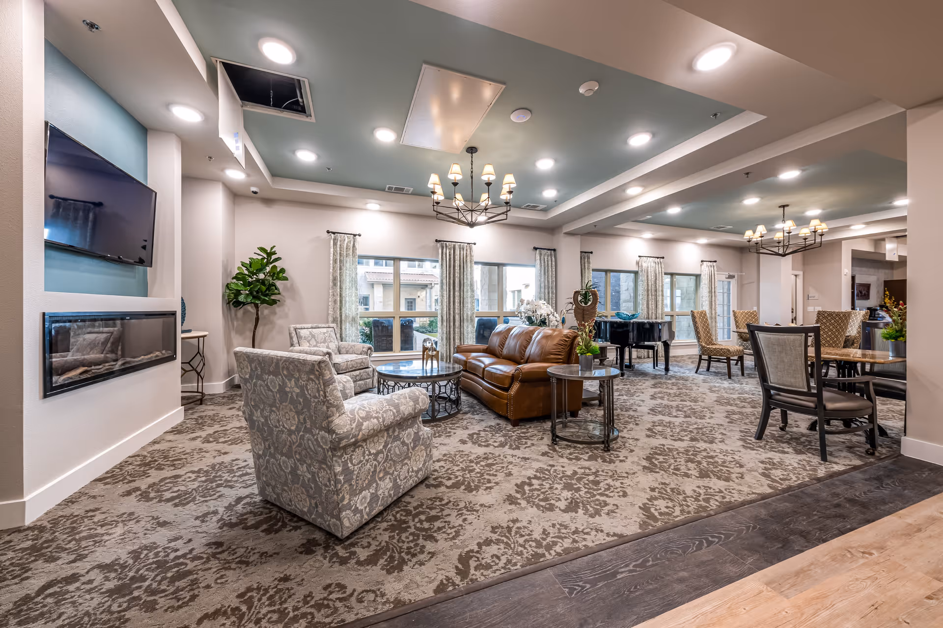 A spacious and well-lit living room area in an assisted living facility featuring a brown leather sofa, patterned armchairs, round coffee tables, a wall-mounted TV above a modern fireplace, large windows with curtains, a grand piano, and multiple dining tables with chairs. The ceiling has recessed lighting and chandeliers, and the floor is covered with a patterned carpet.