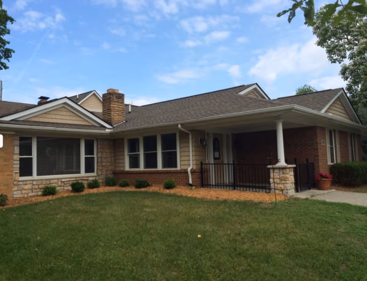 Exterior view of a single-story residential building with a combination of brick, stone, and siding on the facade. The building has a covered porch supported by white columns, several windows, and a well-maintained lawn with small shrubs in front. The sky is partly cloudy.