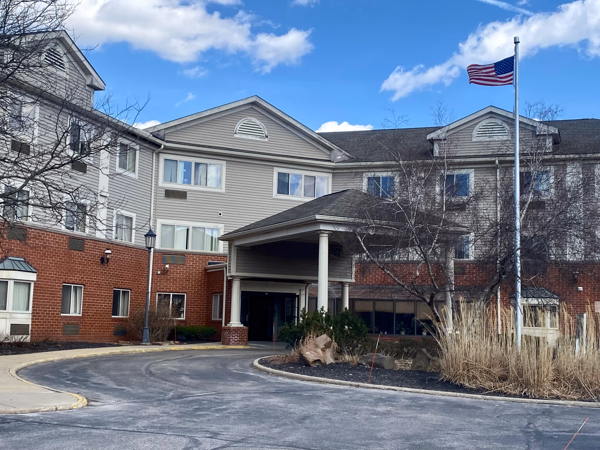 Exterior view of a multi-story senior living facility with a covered entrance, an American flag on a flagpole, and some leafless trees and shrubs in front. The building has a combination of brick and beige siding with several windows.