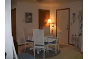 A small dining area with a round table covered by a white tablecloth and four white chairs around it. There is a floor lamp next to a closed wooden door, and a framed picture hanging on the wall above a thermostat. The room has beige walls and carpeted floor.