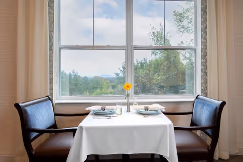 A small dining table set for two with white tablecloth, plates, glasses, and silverware, positioned in front of a large window showing a view of trees and a partly cloudy sky. Two cushioned chairs with wooden armrests are placed on either side of the table.