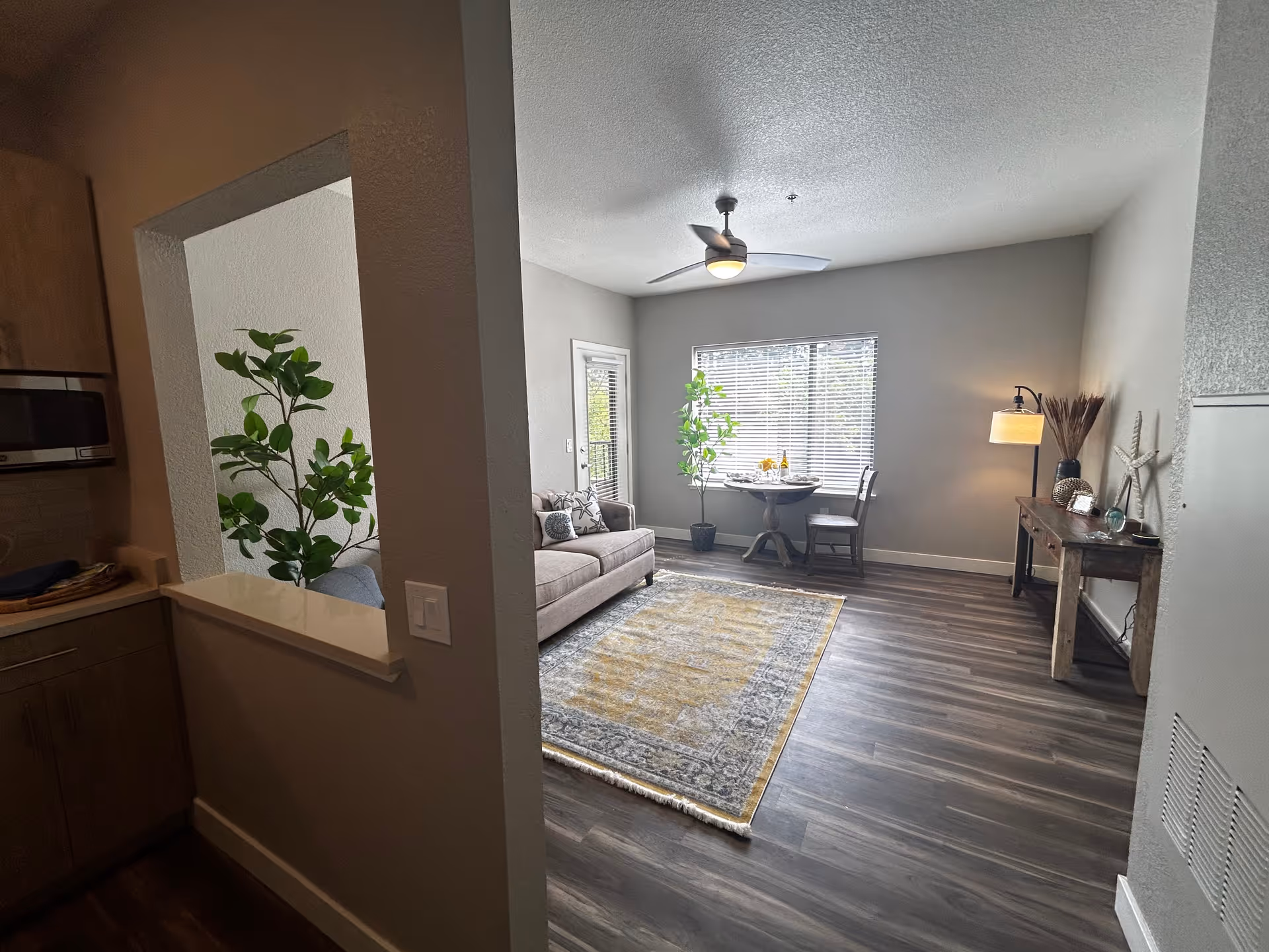 A bright living room with a large window covered by blinds, a ceiling fan with light, a beige sofa with decorative pillows, a small round dining table with two chairs, a potted plant near the window, a wooden console table with a lamp and decorative items, and a patterned area rug on dark wood flooring. The image is taken from the kitchen area, partially visible on the left side.
