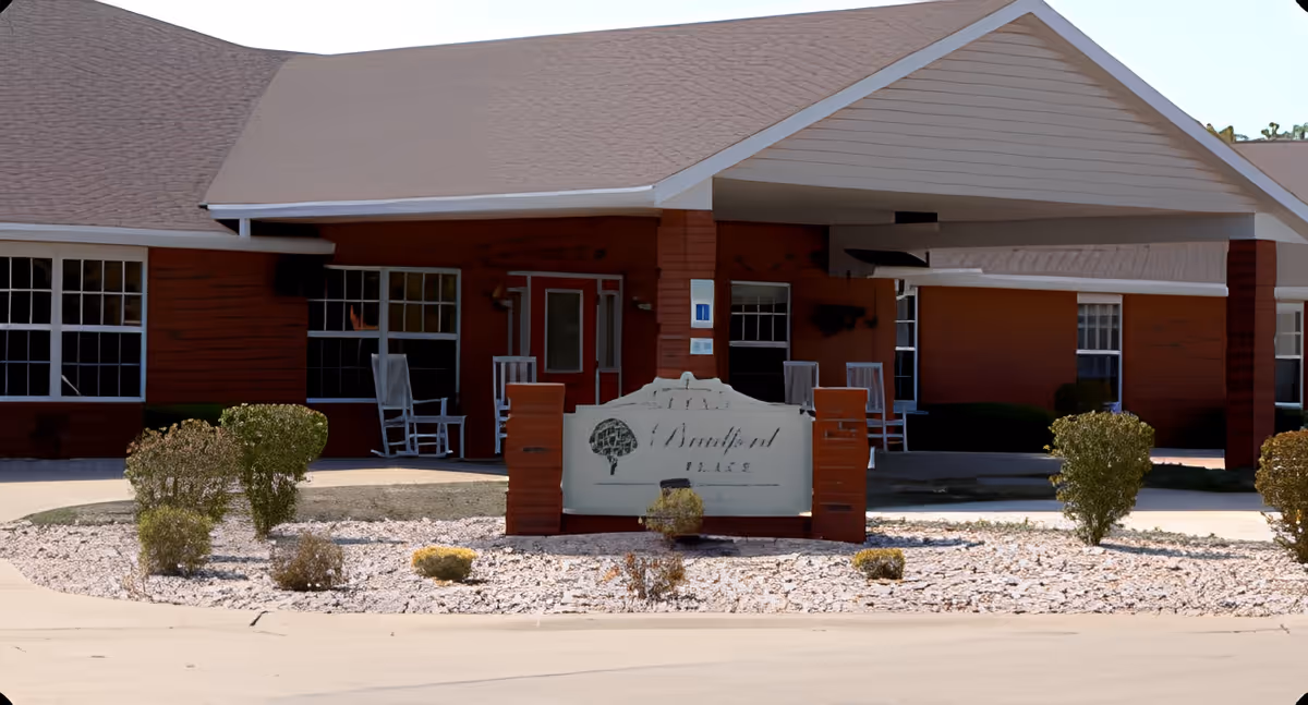 Exterior view of Bradford Place facility showing a single-story building with a covered entrance, several windows, rocking chairs on the porch, and a landscaped area with bushes and rocks in front of a sign displaying the facility name.