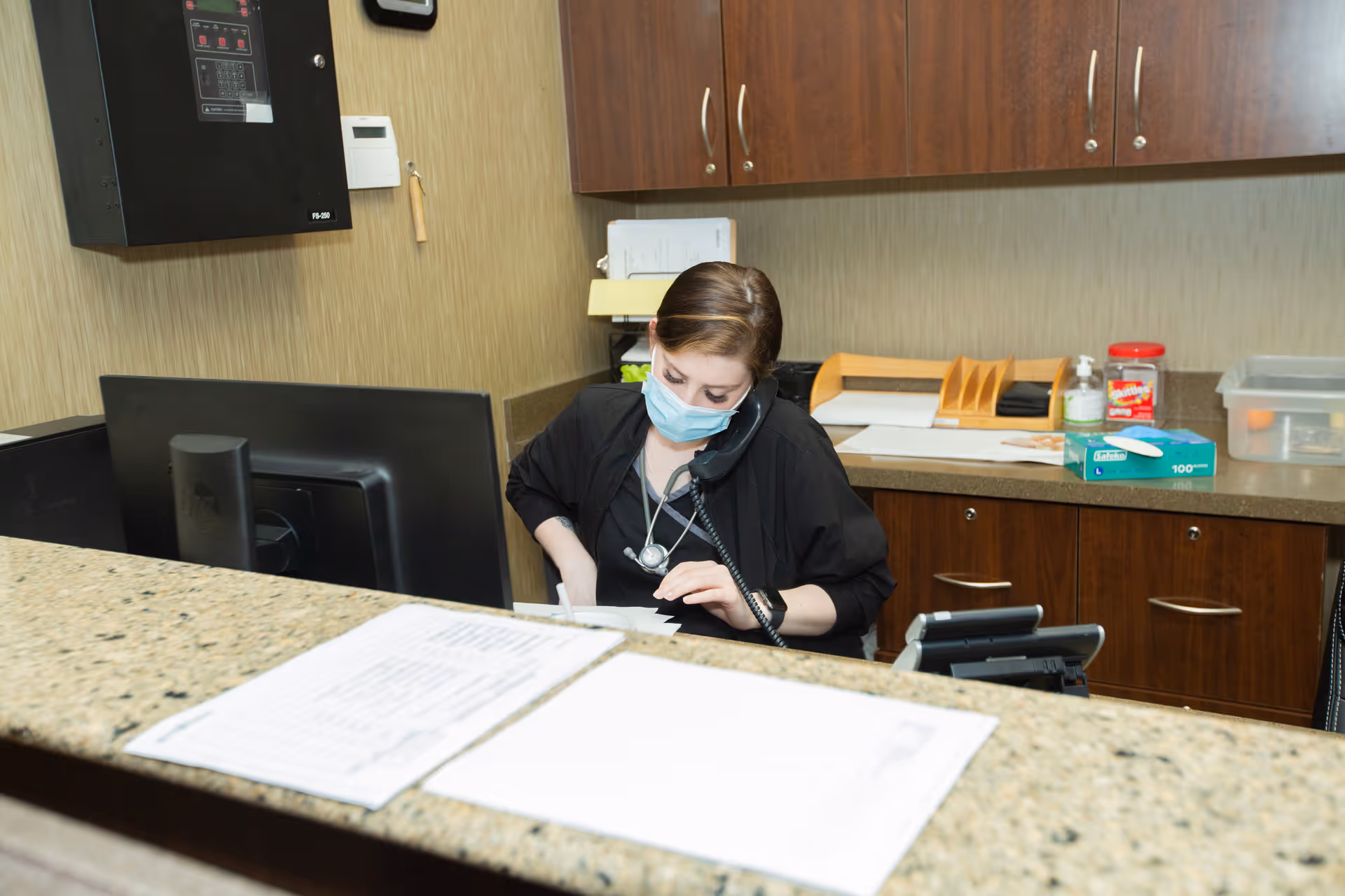 A woman wearing a face mask is sitting behind a reception desk, talking on the phone and writing on a notepad. The desk has a computer monitor and papers on it, and there are cabinets and office supplies in the background.