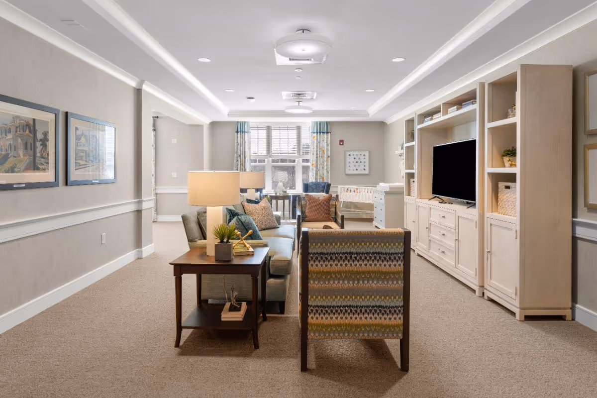 A well-lit living room area in a senior living facility with a sofa, armchair, side table with a lamp and plant, a large built-in entertainment center with a TV, and windows with patterned curtains letting in natural light.