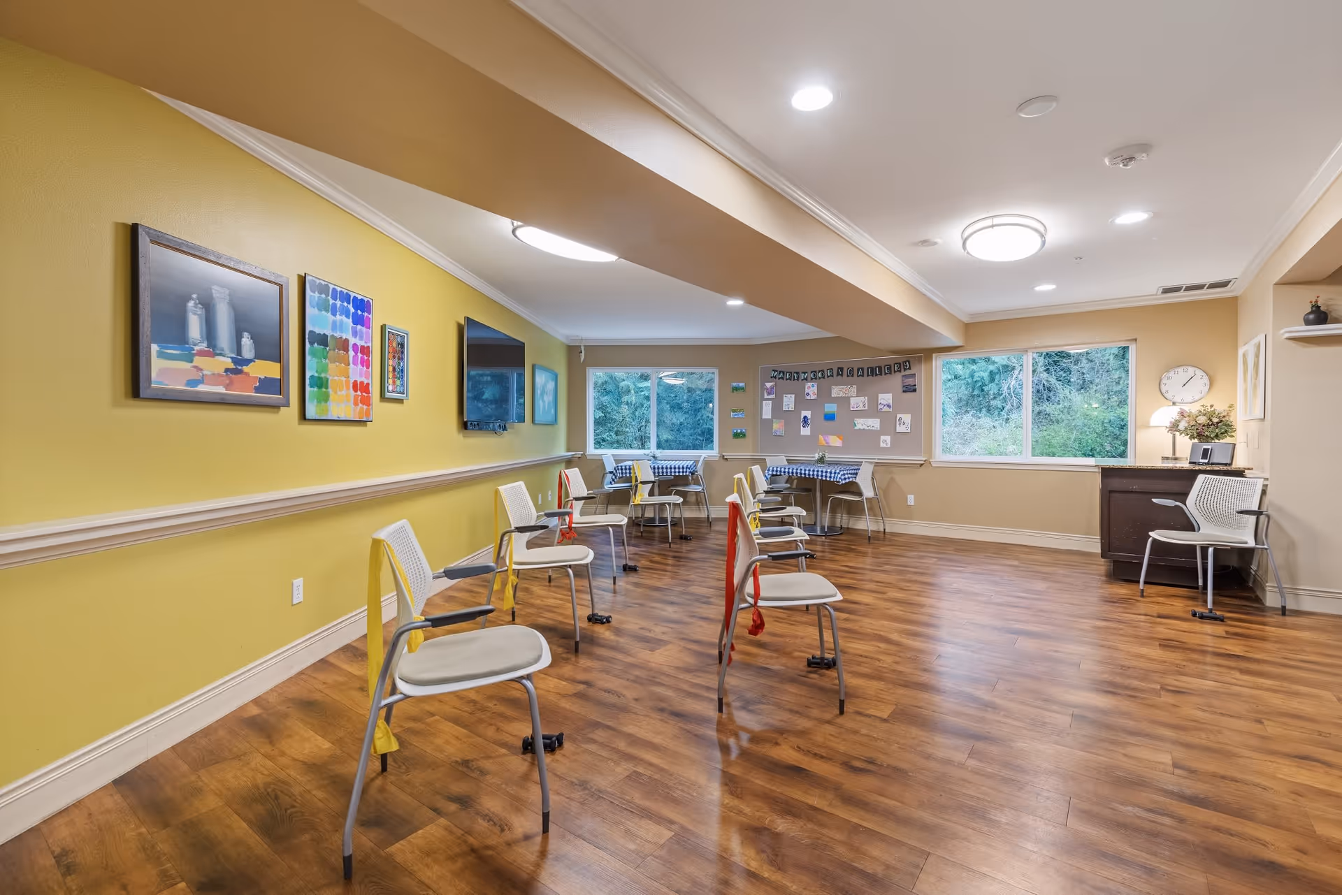 A spacious room with wooden flooring and yellow walls featuring several chairs spaced apart, each with a colored resistance band tied to the backrest. The room has large windows showing greenery outside, a bulletin board with various papers and photos, and a small desk with a clock and a lamp.