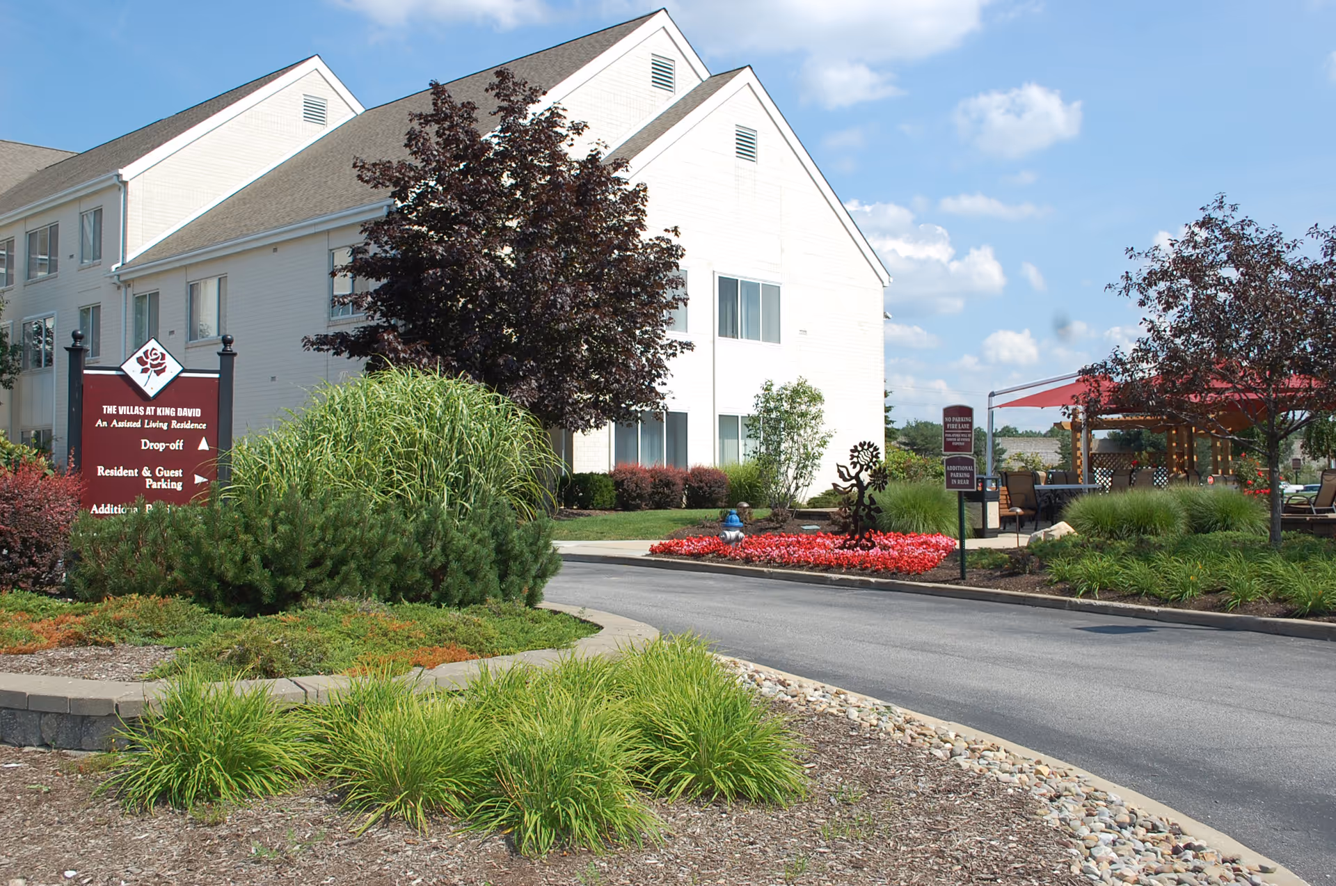 Exterior view of The Villas at King David assisted living residence showing a light-colored building with multiple windows, landscaped greenery including bushes, trees, and flower beds, a paved driveway, and a covered outdoor seating area with red umbrellas under a blue sky with scattered clouds.
