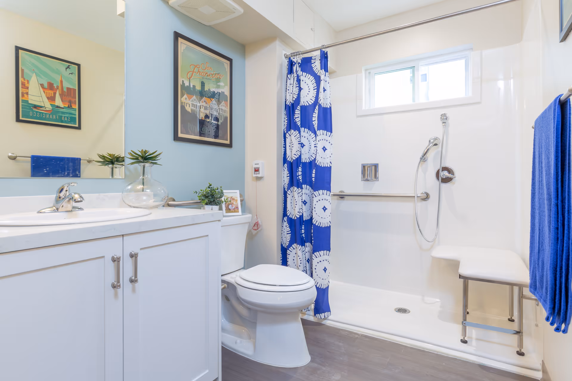 Bright and clean bathroom with a white vanity and sink, a toilet, and a walk-in shower with a blue and white patterned shower curtain. The shower has a handheld showerhead, a grab bar, and a foldable bench. There are two framed pictures on the wall, a small window above the shower, and blue towels hanging on a towel rack.