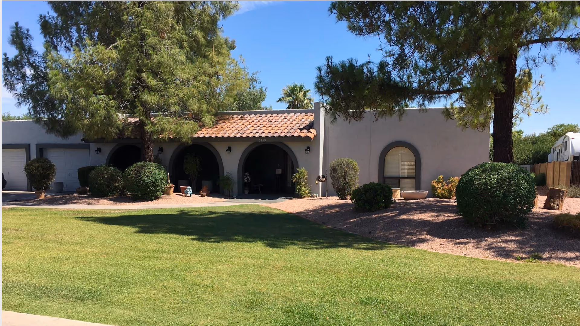 Single-story building with a tiled roof and arched entryways surrounded by trees, bushes, and a well-maintained lawn under a clear blue sky.