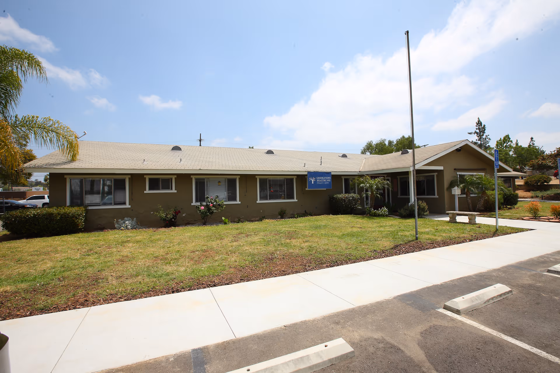 Single-story building with a beige exterior and a light-colored roof, surrounded by a grassy lawn and some small bushes. There is a sidewalk and parking spaces in front of the building, along with a flagpole and a sign on the building that reads 'Generations Healthcare'. The sky is partly cloudy.