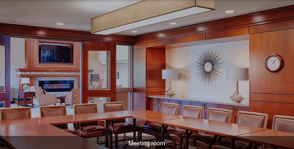 A meeting room with a large U-shaped wooden table surrounded by brown leather chairs. The room features wood-paneled walls, two table lamps with white shades on a granite countertop, and a decorative sunburst wall clock. Through glass doors, a cozy lounge area with armchairs, a fireplace, and a mounted TV is visible.