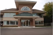 Front exterior view of a two-story building with large windows and a covered entrance with a green roof, surrounded by trees.