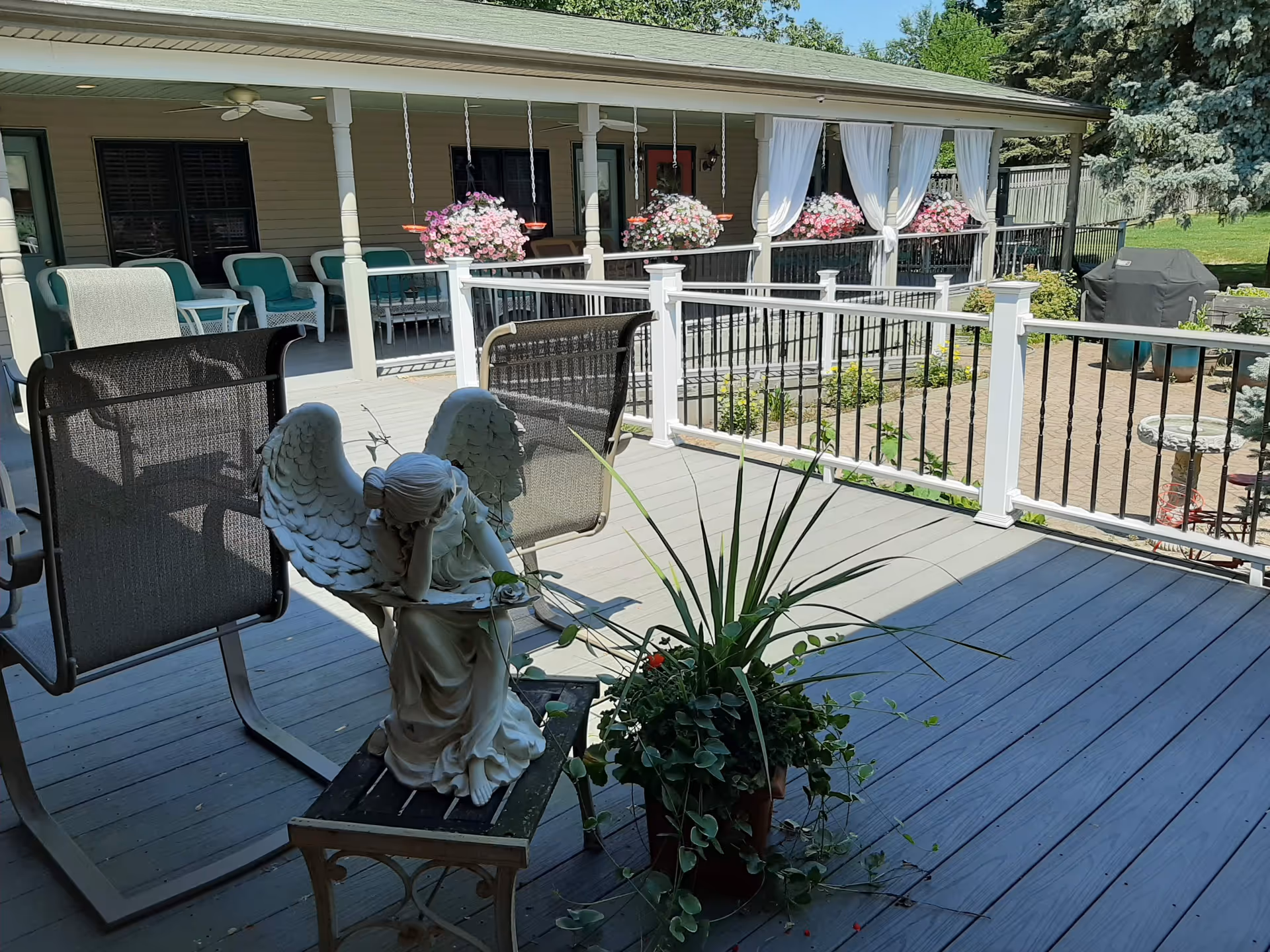 Outdoor patio area with a wooden deck featuring a white angel statue sitting on a small table, surrounded by potted plants. There are several chairs and a covered porch with hanging flower baskets and white curtains. A grill and additional seating are visible in the background along with green trees and a lawn.