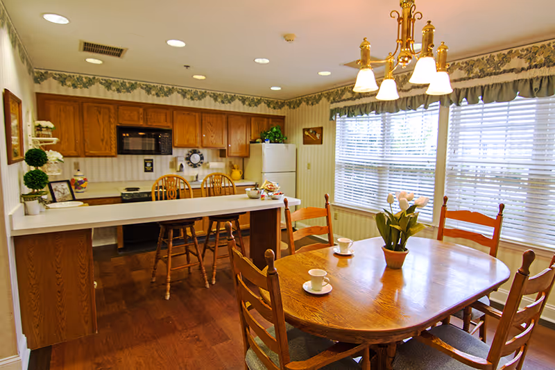 A bright kitchen and dining area with wooden cabinets, a white refrigerator, a microwave, and a kitchen island with two wooden chairs. The dining table has four wooden chairs and is set with cups and saucers, with a potted plant centerpiece. Large windows with blinds allow natural light to fill the room, and a chandelier hangs above the dining table.