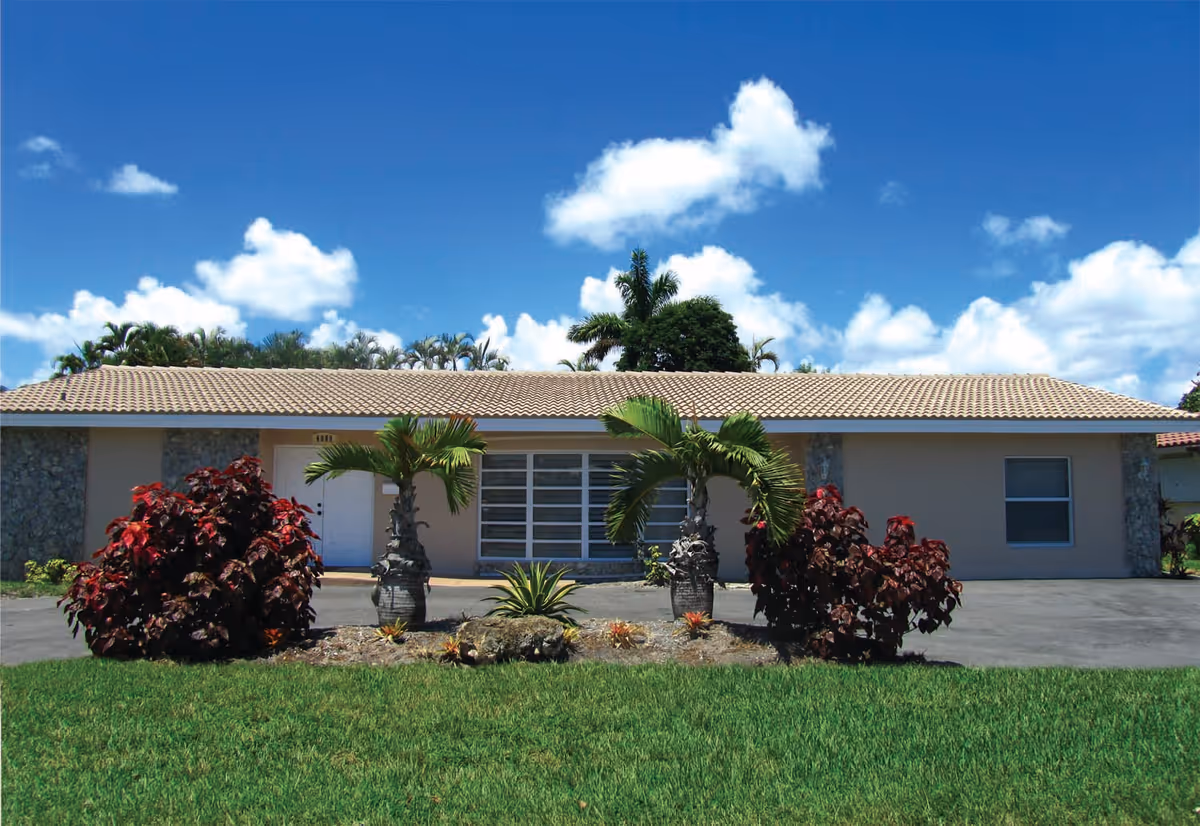 Single-story building with a tiled roof, beige walls, and a white door, surrounded by green grass, palm trees, and red-leafed bushes under a blue sky with scattered clouds.