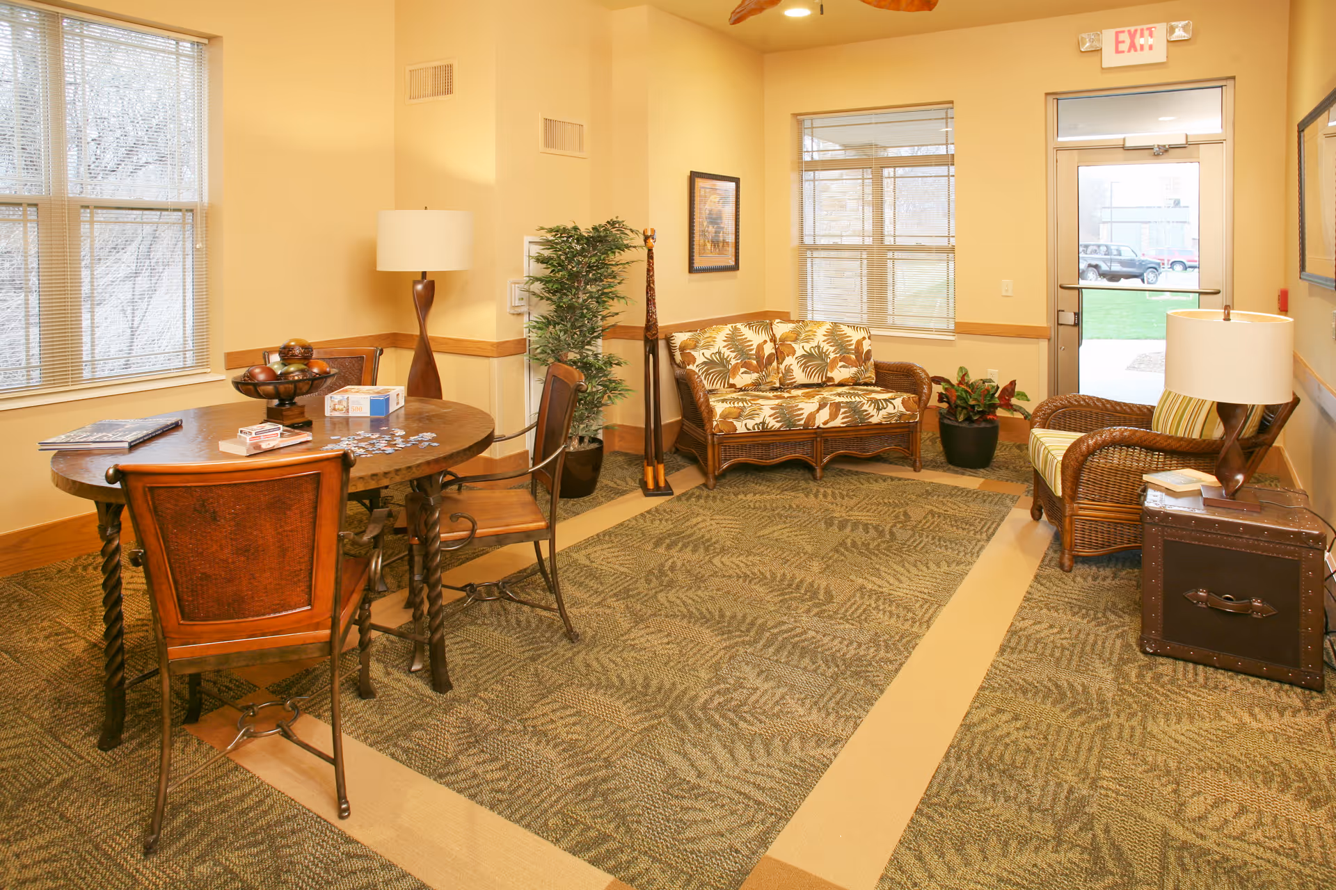 A cozy sitting area in a senior living facility with a round wooden table and four chairs on the left side, a wicker loveseat with floral cushions against the back wall, a wicker armchair with a striped cushion on the right, two potted plants, a floor lamp, and a side table with a lamp and books. Large windows and a glass door provide natural light.