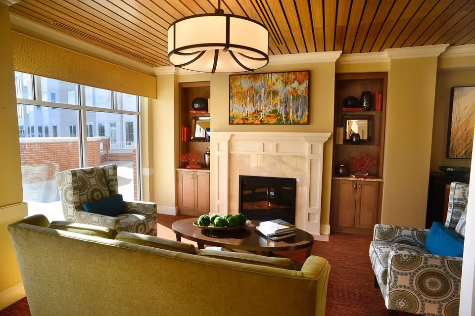 A cozy living room area with a green sofa and two patterned armchairs with blue cushions arranged around a wooden coffee table. The room features a white fireplace with a colorful painting above it, built-in wooden shelves on either side, and a large window letting in natural light. The ceiling has wooden paneling and a large round light fixture.
