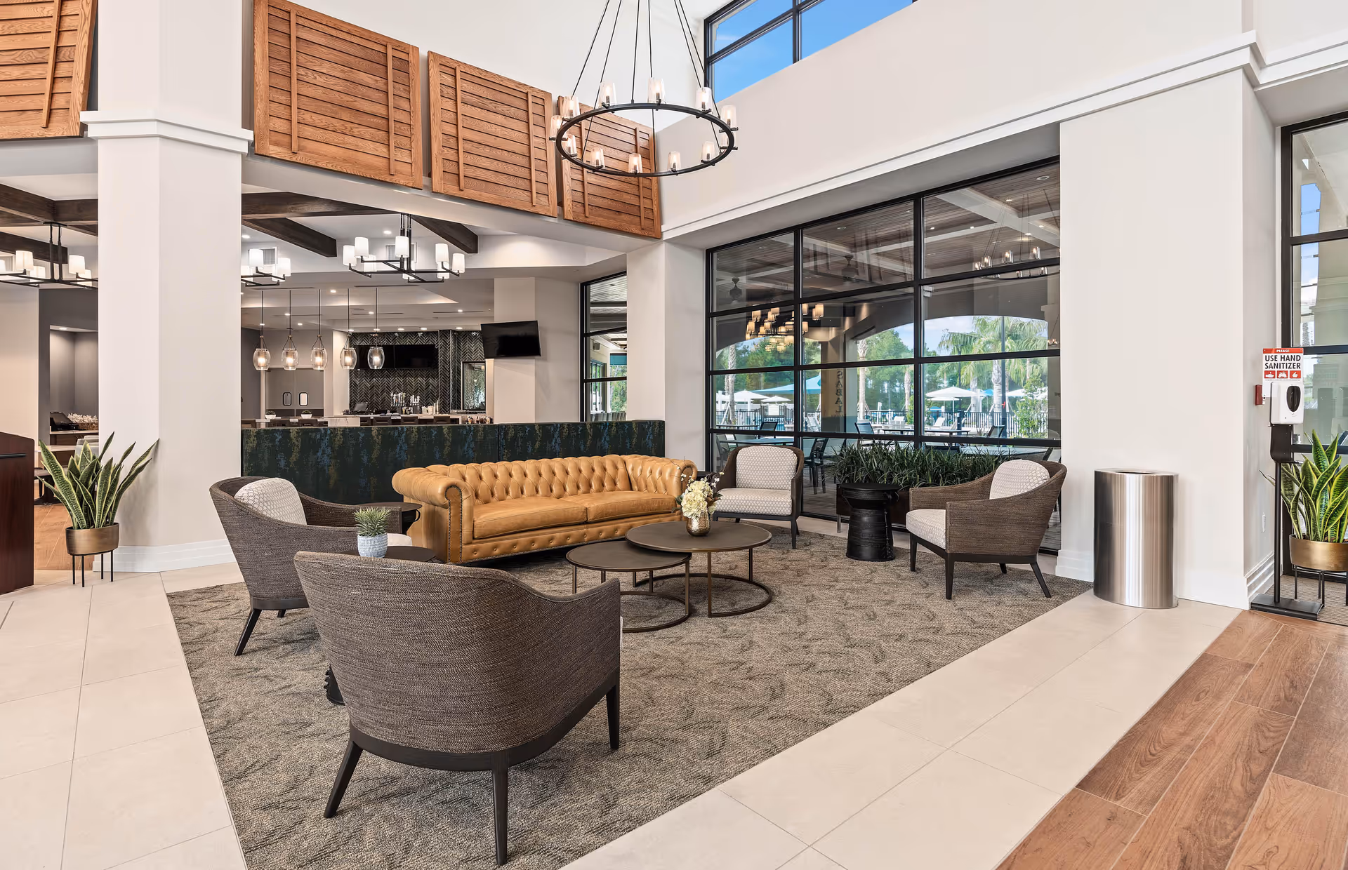 A spacious and modern living room area in a senior living facility featuring a brown leather sofa, four gray armchairs arranged around two round coffee tables, large windows letting in natural light, indoor plants, and contemporary light fixtures hanging from the ceiling.