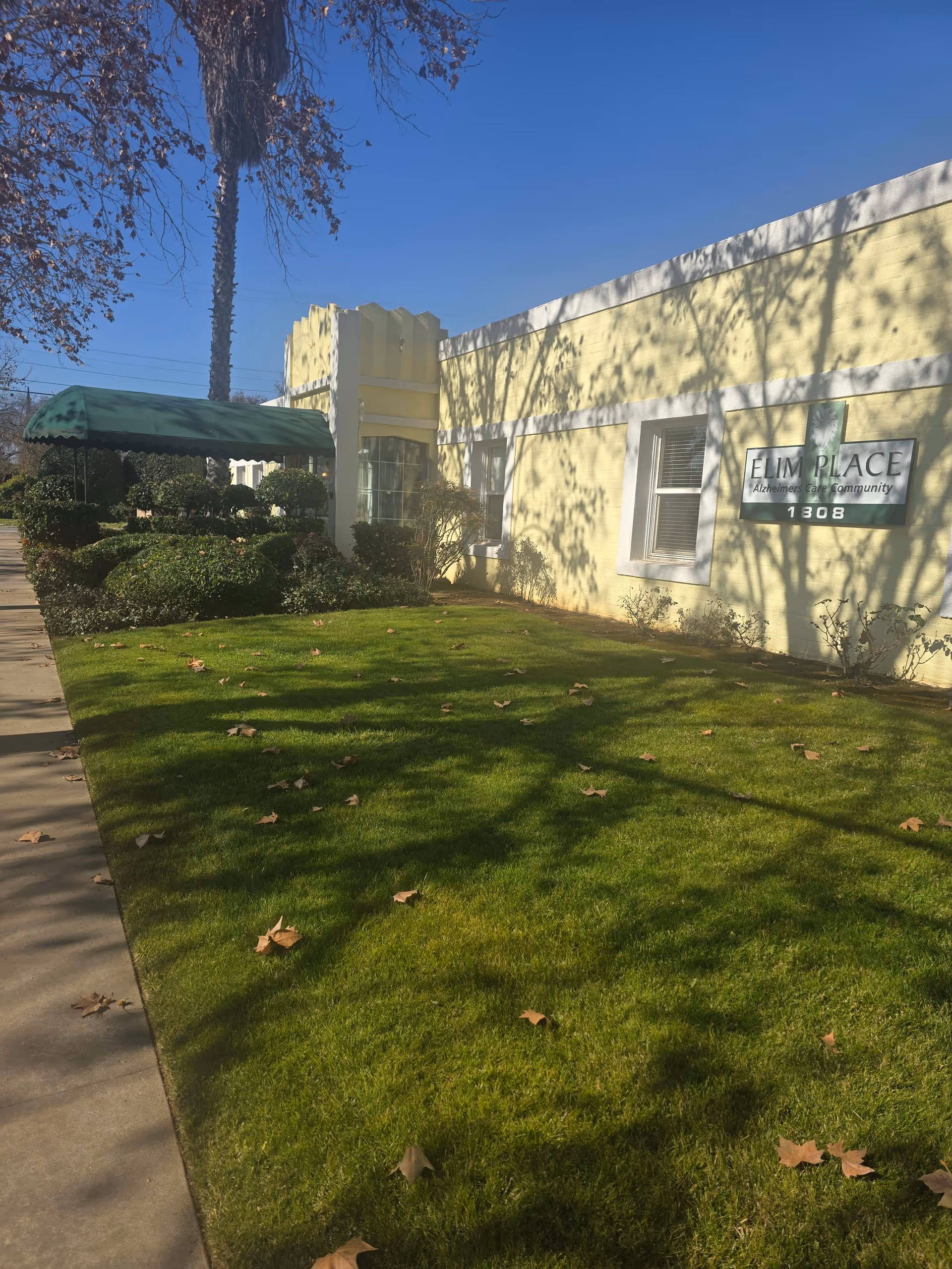 Exterior view of a yellow building with a green awning over a walkway, surrounded by trimmed bushes and a green lawn with scattered fallen leaves under a clear blue sky. A sign on the building reads 'Elim Place Alzheimer's Care Community 1308'.