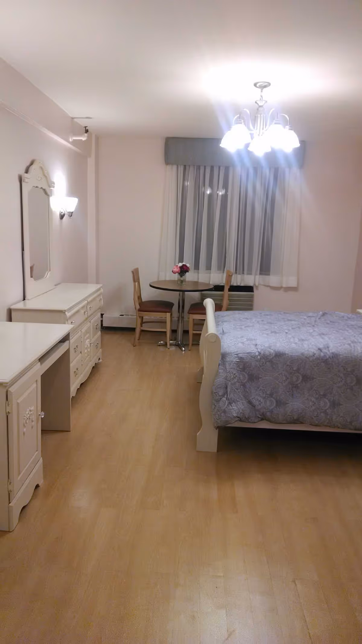 A senior living facility bedroom with a bed covered in a gray patterned comforter, a small round table with two chairs near a window with sheer curtains, a white dresser with a mirror, and a chandelier light fixture on the ceiling.