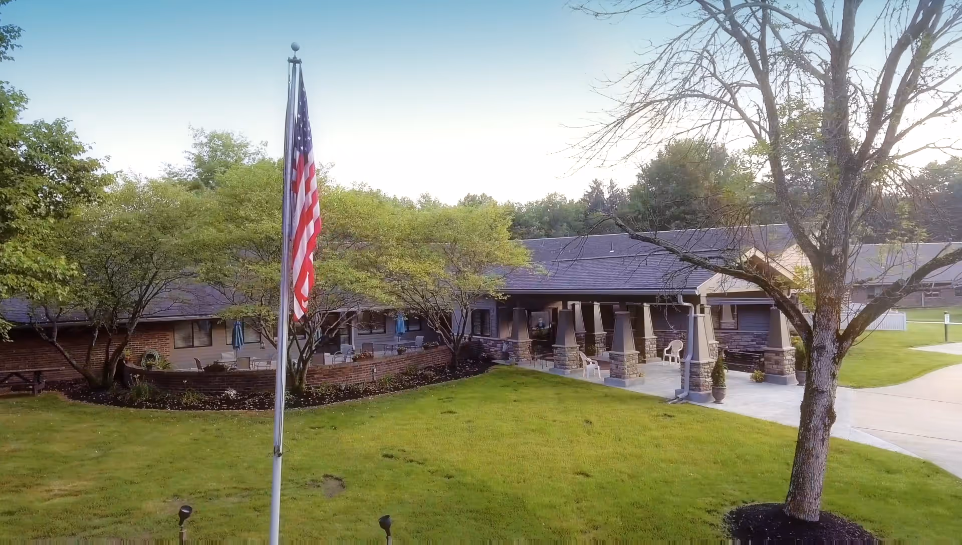 Exterior view of Warren Manor facility showing a single-story building with a covered entrance supported by stone pillars, surrounded by green lawn, trees, and an American flag on a flagpole in the foreground.