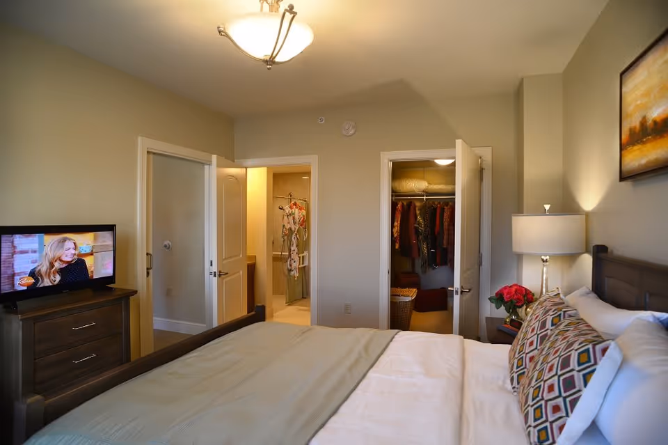 A cozy bedroom with a neatly made bed featuring patterned pillows and a beige blanket. To the right of the bed is a nightstand with a lamp and a vase of red flowers. On the left side of the room, there is a wooden dresser with a television showing a woman on screen. Two open doors reveal a walk-in closet with hanging clothes and a bathroom with a shower and hanging garments. The walls are painted light beige, and a framed painting hangs above the bed.