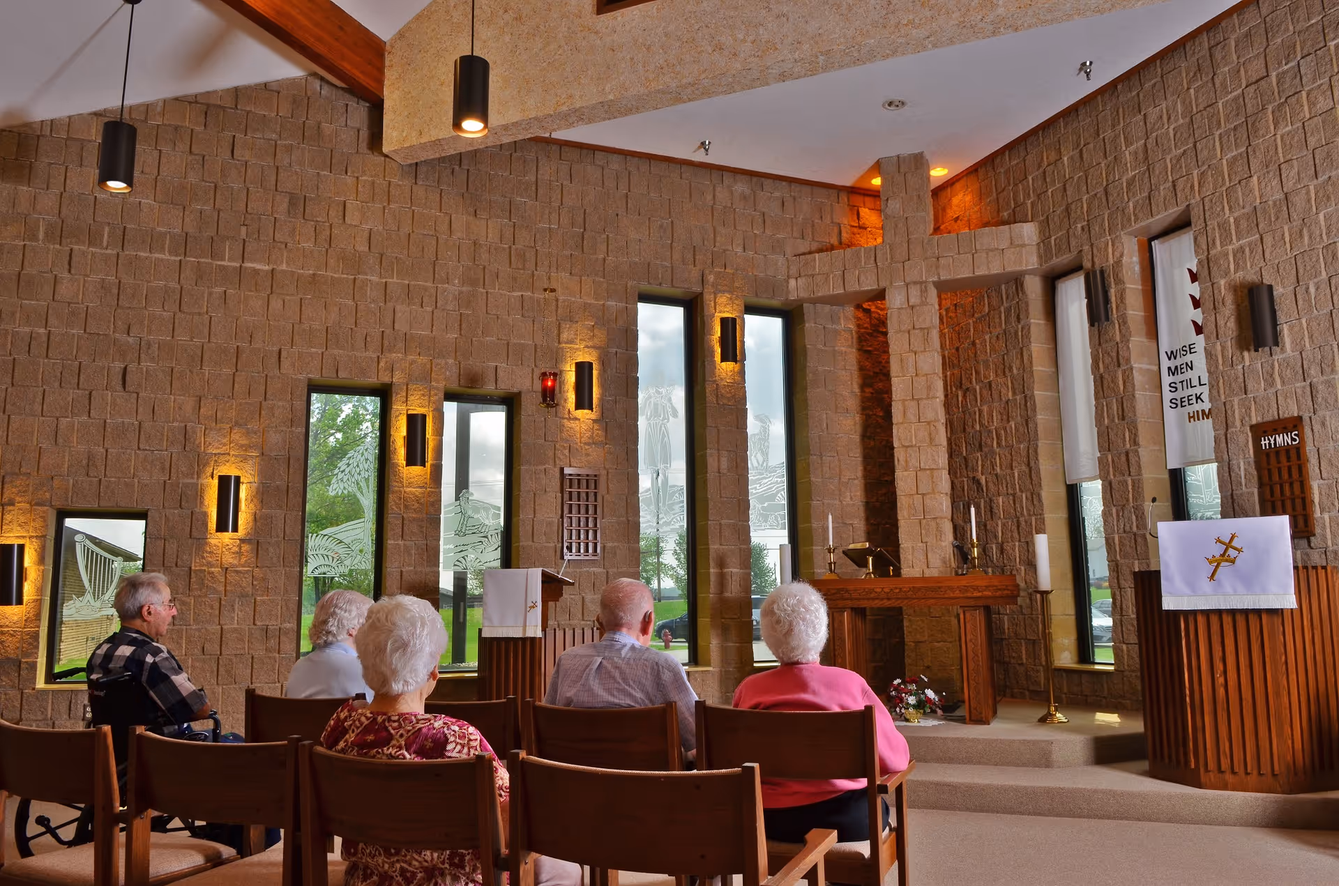 Interior of a chapel with stone walls and tall narrow windows. Five elderly people are seated on wooden chairs facing the altar, which features a large stone cross, candles, and religious banners on the walls.