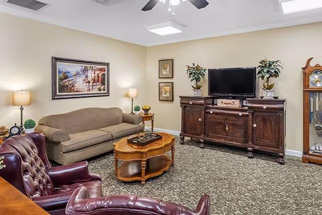 A cozy living room with a patterned carpet, a beige sofa, two burgundy leather armchairs, a wooden coffee table, a dark wooden TV cabinet with a flat-screen TV, two potted plants, a grandfather clock, and framed artwork on the walls.