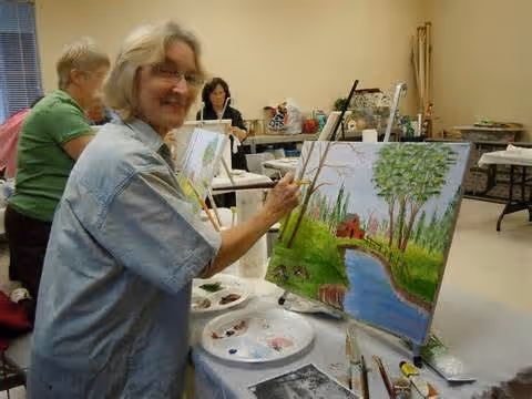 A group of elderly women engaged in a painting activity in a well-lit room. One woman in the foreground is smiling at the camera while working on a landscape painting featuring a river, trees, and greenery. Other women are seated at tables in the background, also painting.