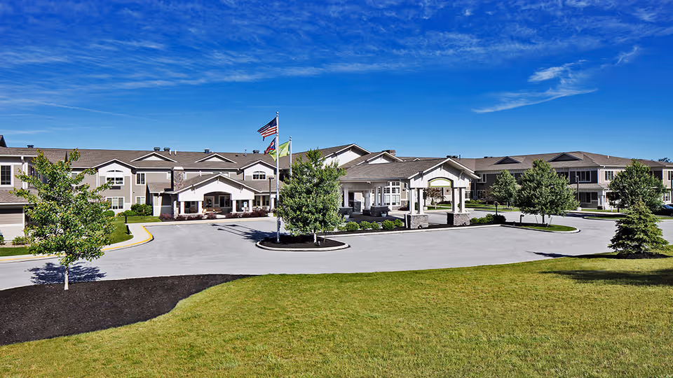 Exterior view of The Enclave of Newell Creek senior living facility on a clear sunny day, showing a large driveway, landscaped green lawn, several trees, and the building with multiple windows and a covered entrance with flags flying.