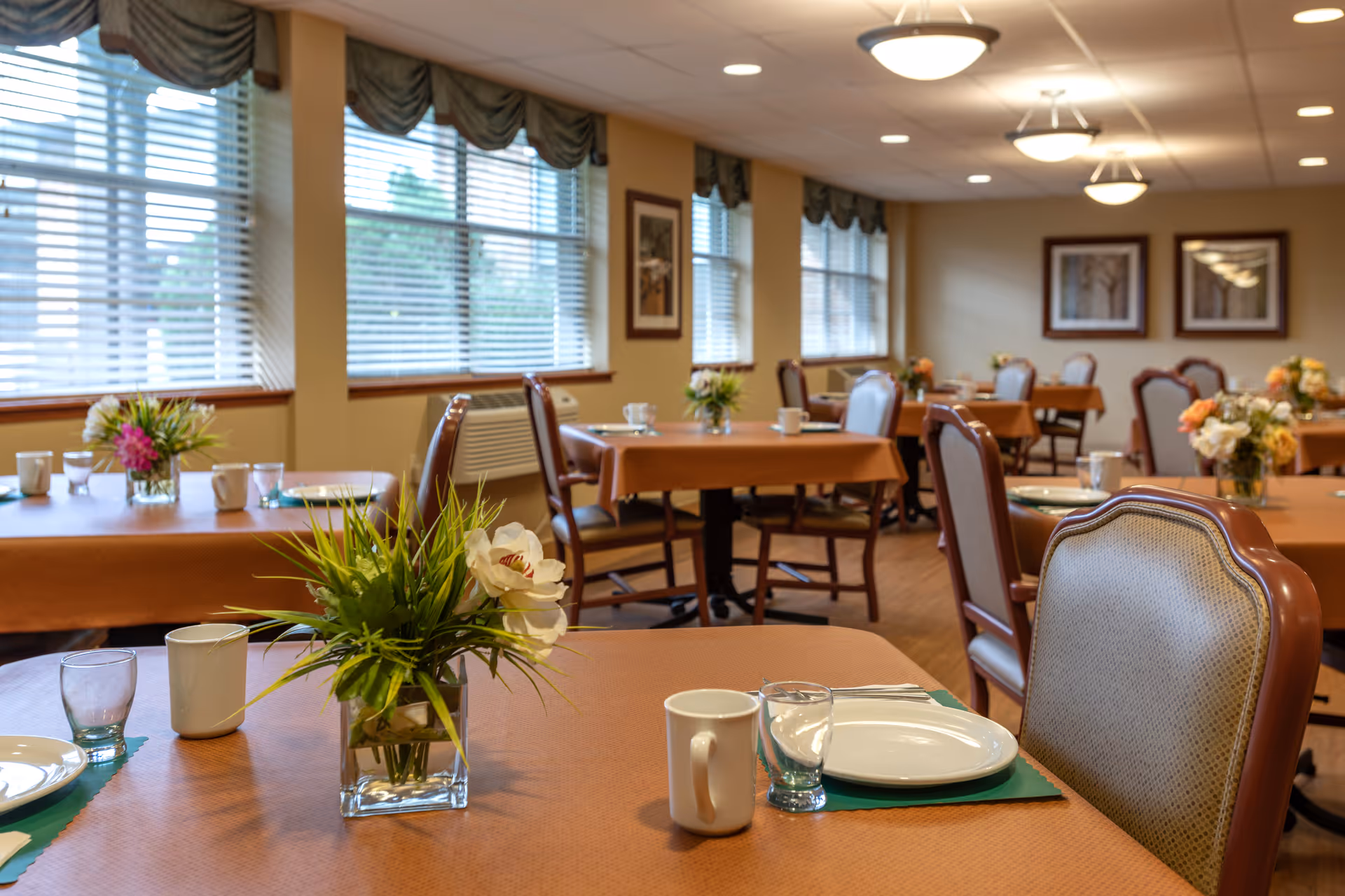 A dining room in a senior living facility with multiple tables covered in brown tablecloths, each set with white plates, cups, glasses, and floral centerpieces. The room has large windows with blinds and framed artwork on the walls, and is softly lit by ceiling lights.