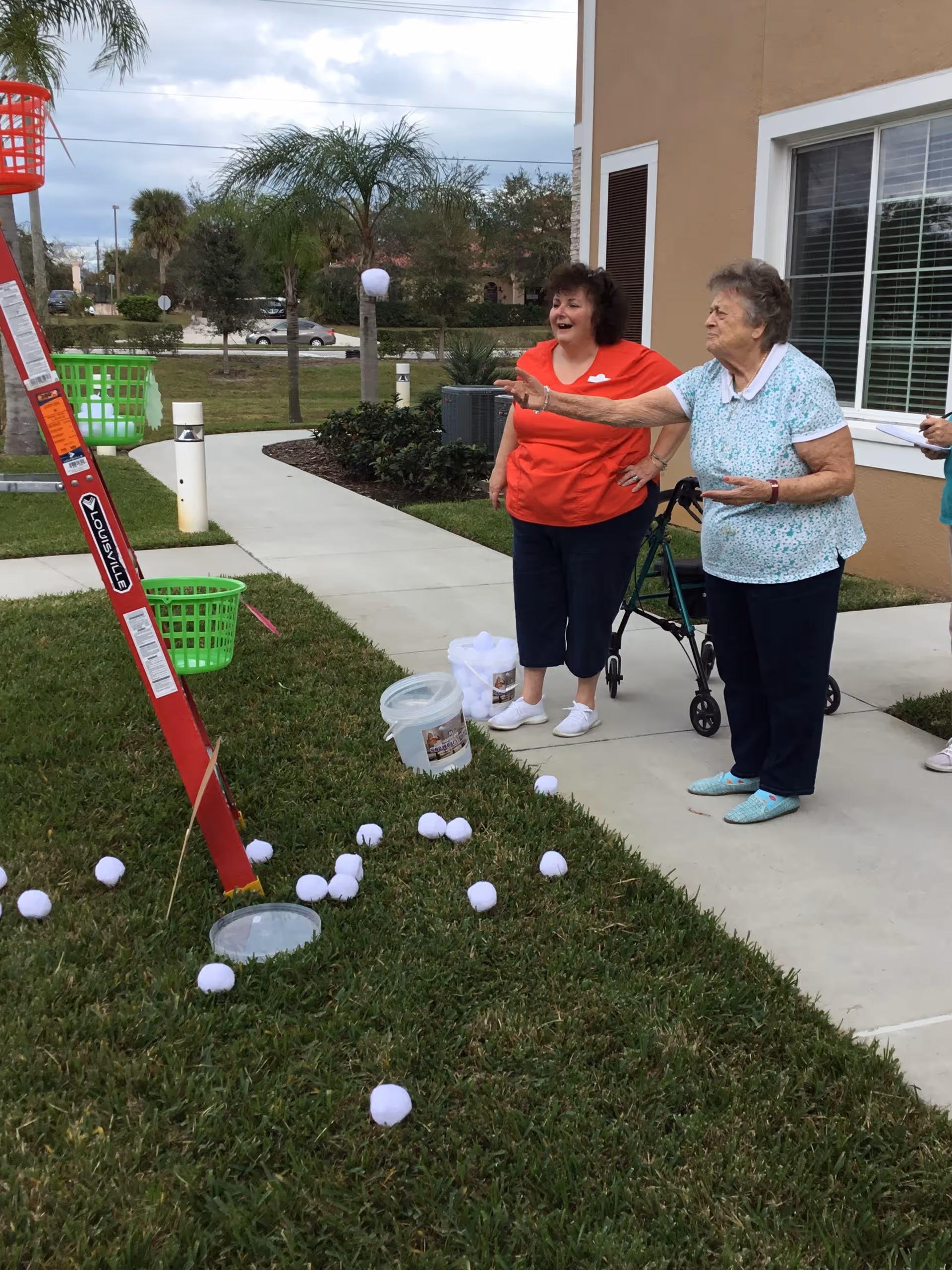 Two elderly women playing an outdoor game involving tossing white balls into baskets attached to a red ladder. One woman is in a red shirt and black pants, smiling, while the other woman in a light blue patterned shirt and dark pants is in the act of throwing a ball. They are standing on a sidewalk next to a grassy area with scattered white balls and buckets. A walker is visible behind the woman throwing the ball, and a beige building with a window is in the background.