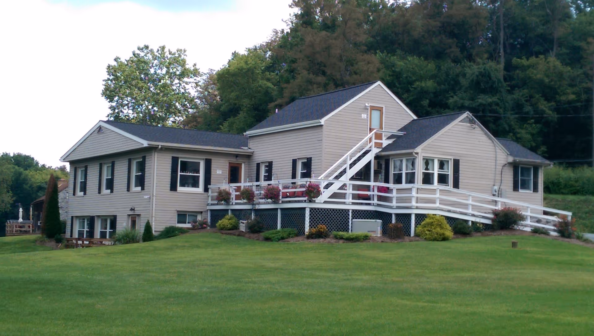 A beige two-story house with black shutters and a black roof, surrounded by a well-maintained green lawn and trees in the background. The house features a white railing along a porch and ramp, with flower pots placed on the railing.
