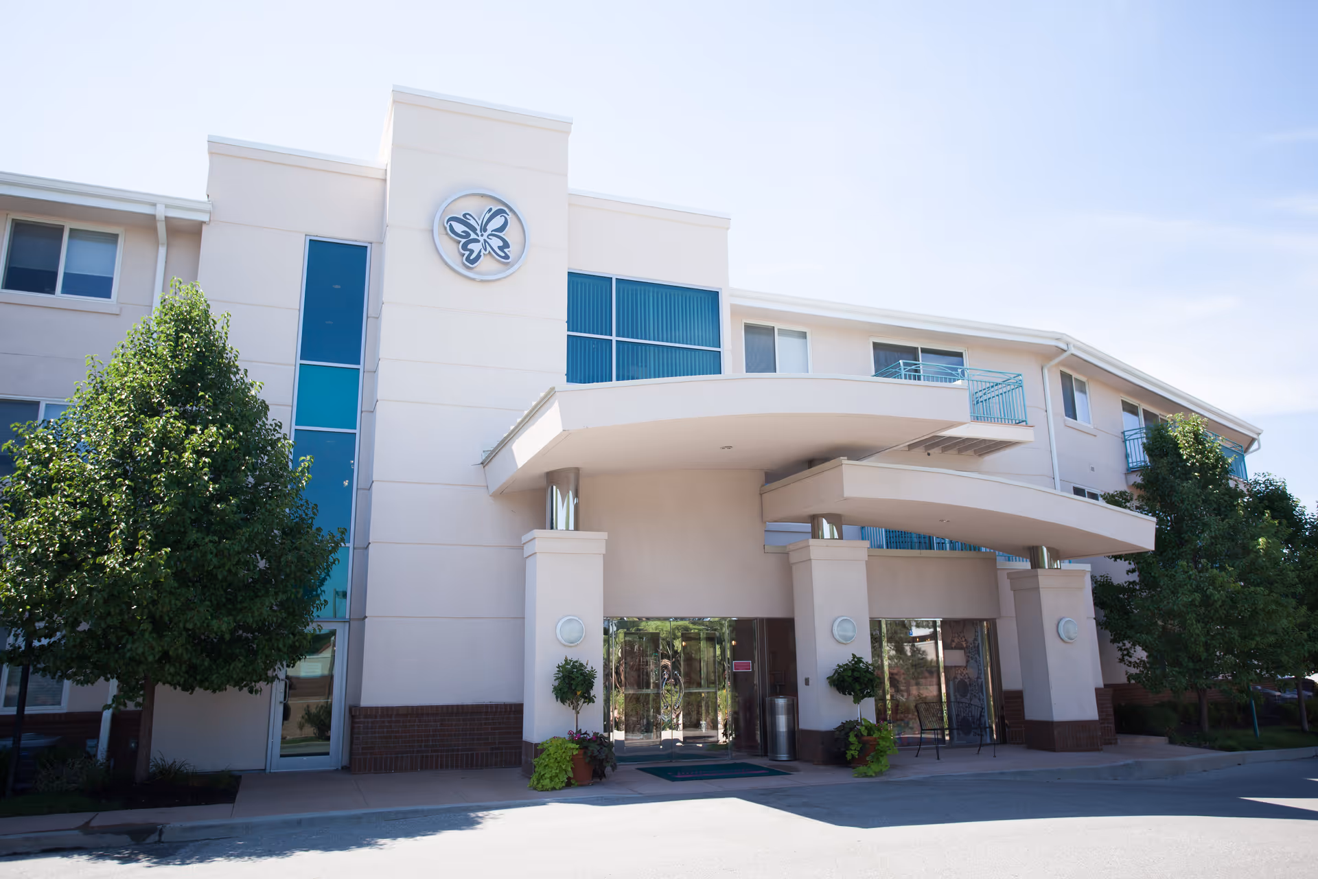 Front exterior view of a multi-story senior living facility building with a covered entrance, glass doors, potted plants, and trees on either side under a clear blue sky.