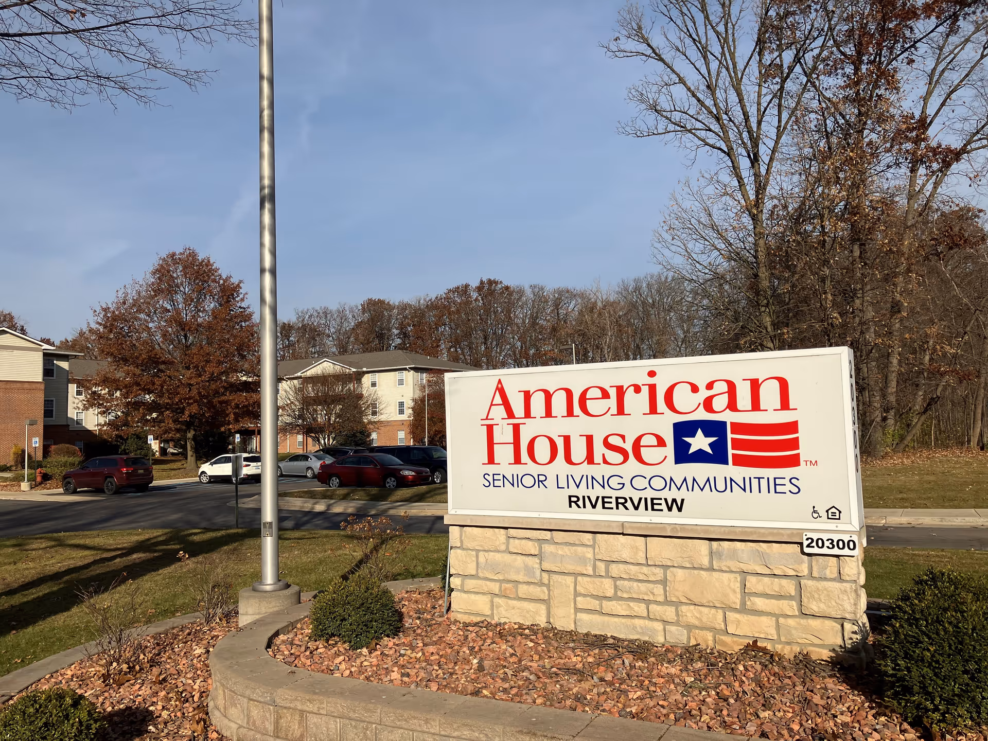 Outdoor view of the entrance sign for American House Senior Living Communities Riverview, with residential buildings, parked cars, trees with autumn leaves, and a clear sky in the background.