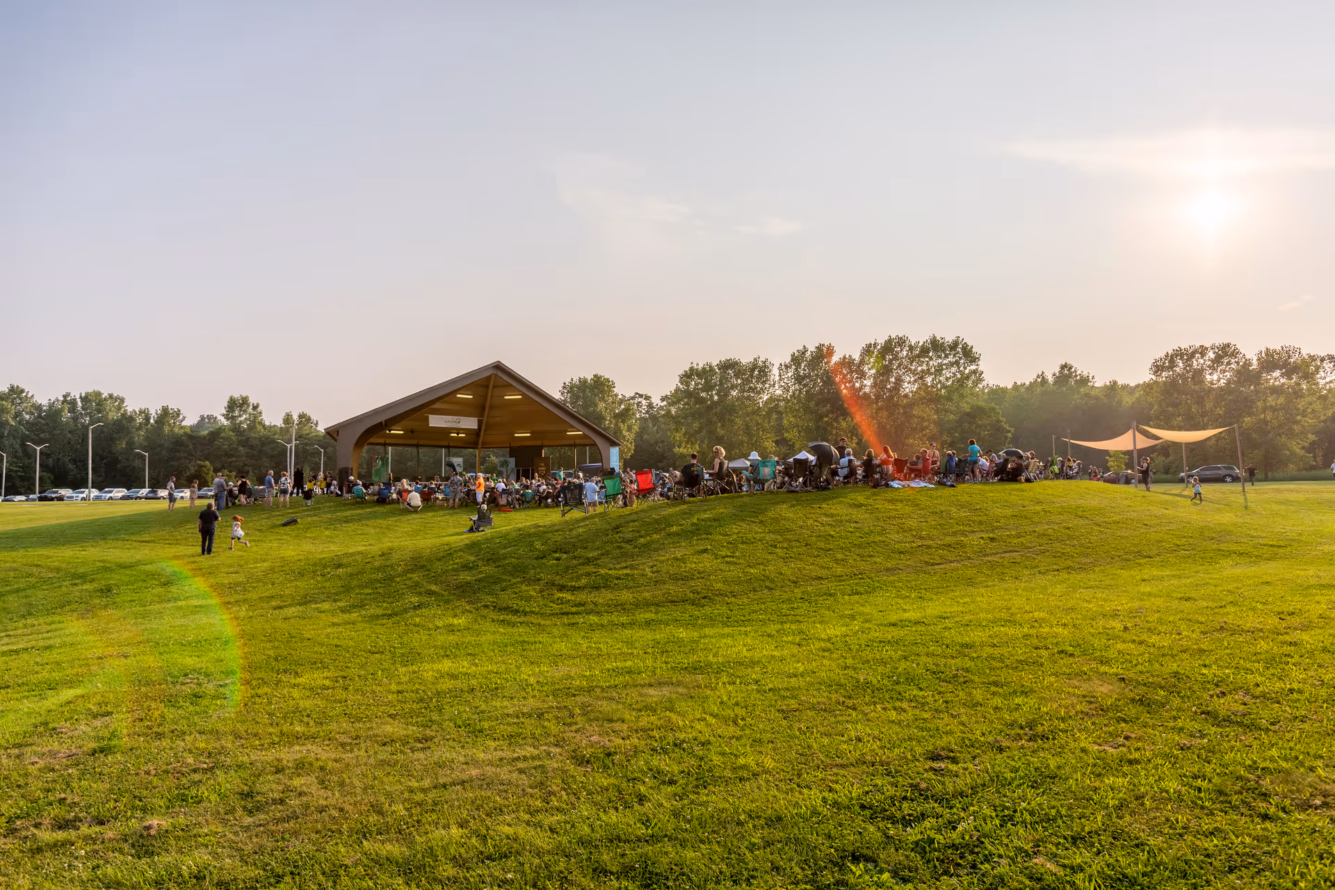 A large group of people gathered outdoors on a grassy hill near a pavilion with a peaked roof. The scene is set in the late afternoon with the sun low in the sky, casting a warm light. Trees line the background and there are some cars parked in the distance.