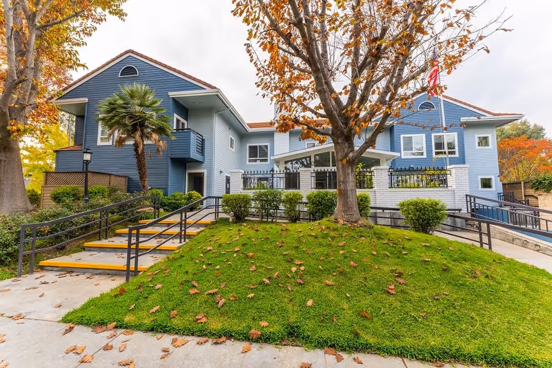 Exterior view of a two-story blue building with white trim, surrounded by green grass and trees with autumn leaves. There are stairs and ramps with black railings leading up to the entrance, which is partially obscured by a tree in the foreground. The sky is overcast.