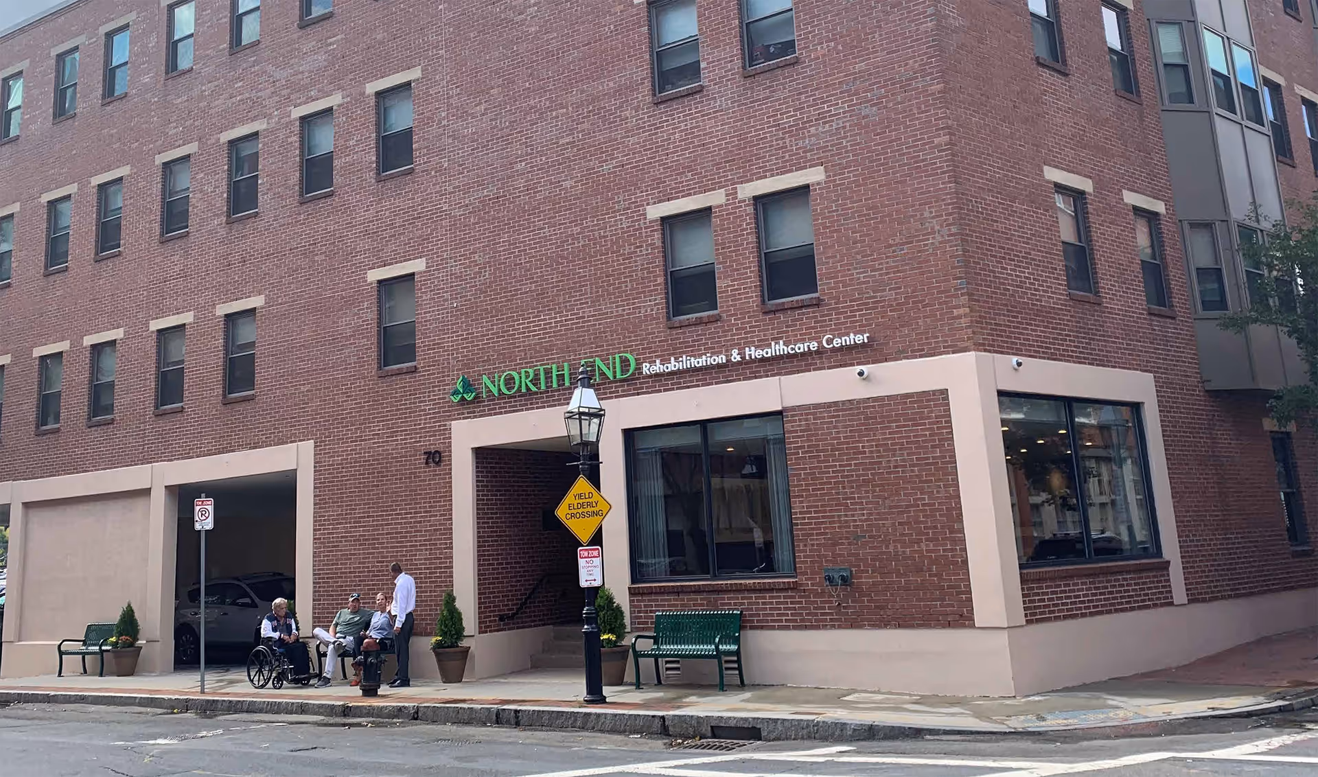 Exterior view of a multi-story brick building with the sign 'North End Rehabilitation & Healthcare Center'. There are four people outside near the entrance, including one person in a wheelchair. A street lamp with a yellow 'Yield Elderly Crossing' sign is visible, along with benches and potted plants on the sidewalk.