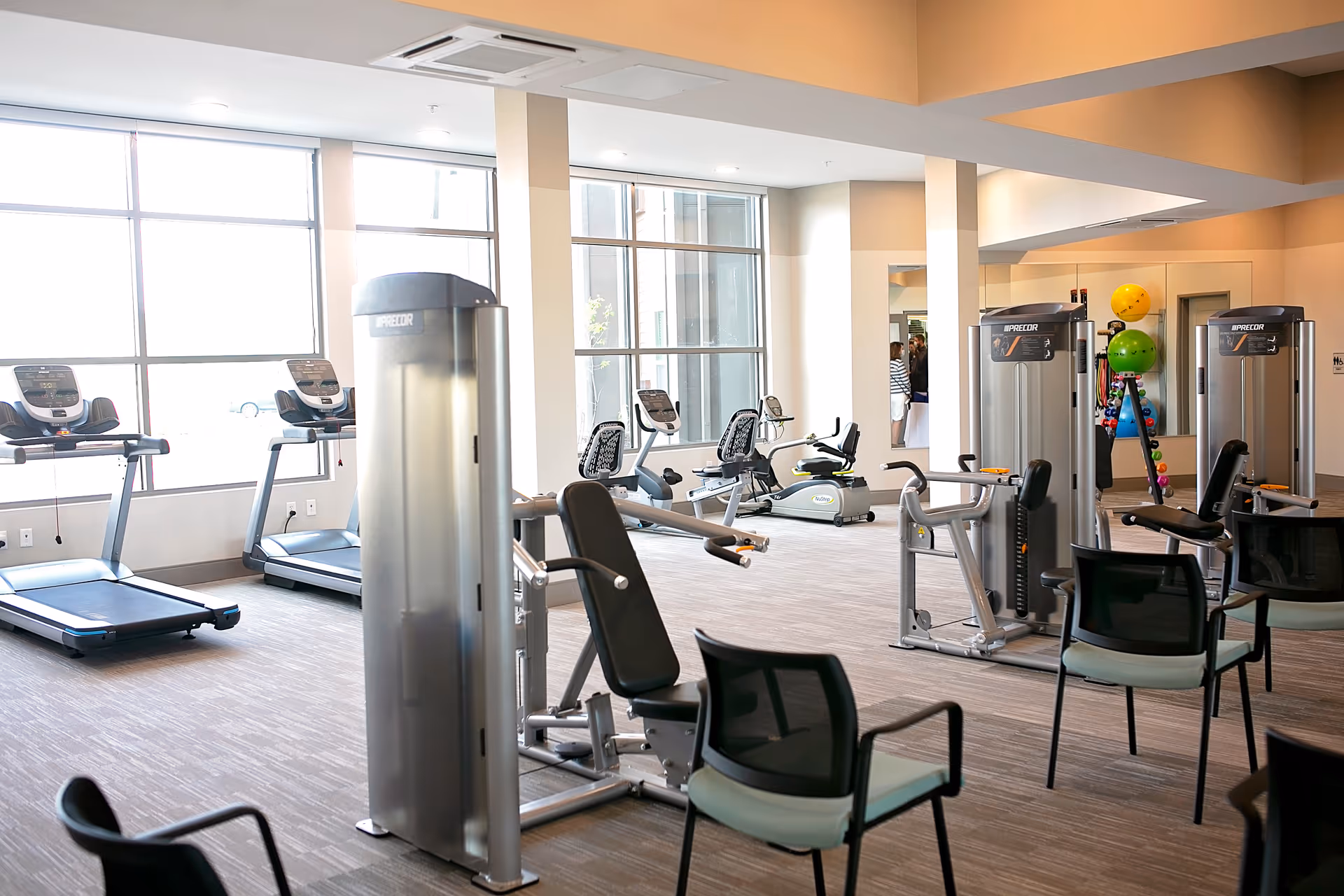 A bright and spacious fitness room with large windows letting in natural light. The room contains various exercise equipment including treadmills, stationary bikes, and strength training machines. There are also chairs placed around the room and colorful exercise balls in the background.