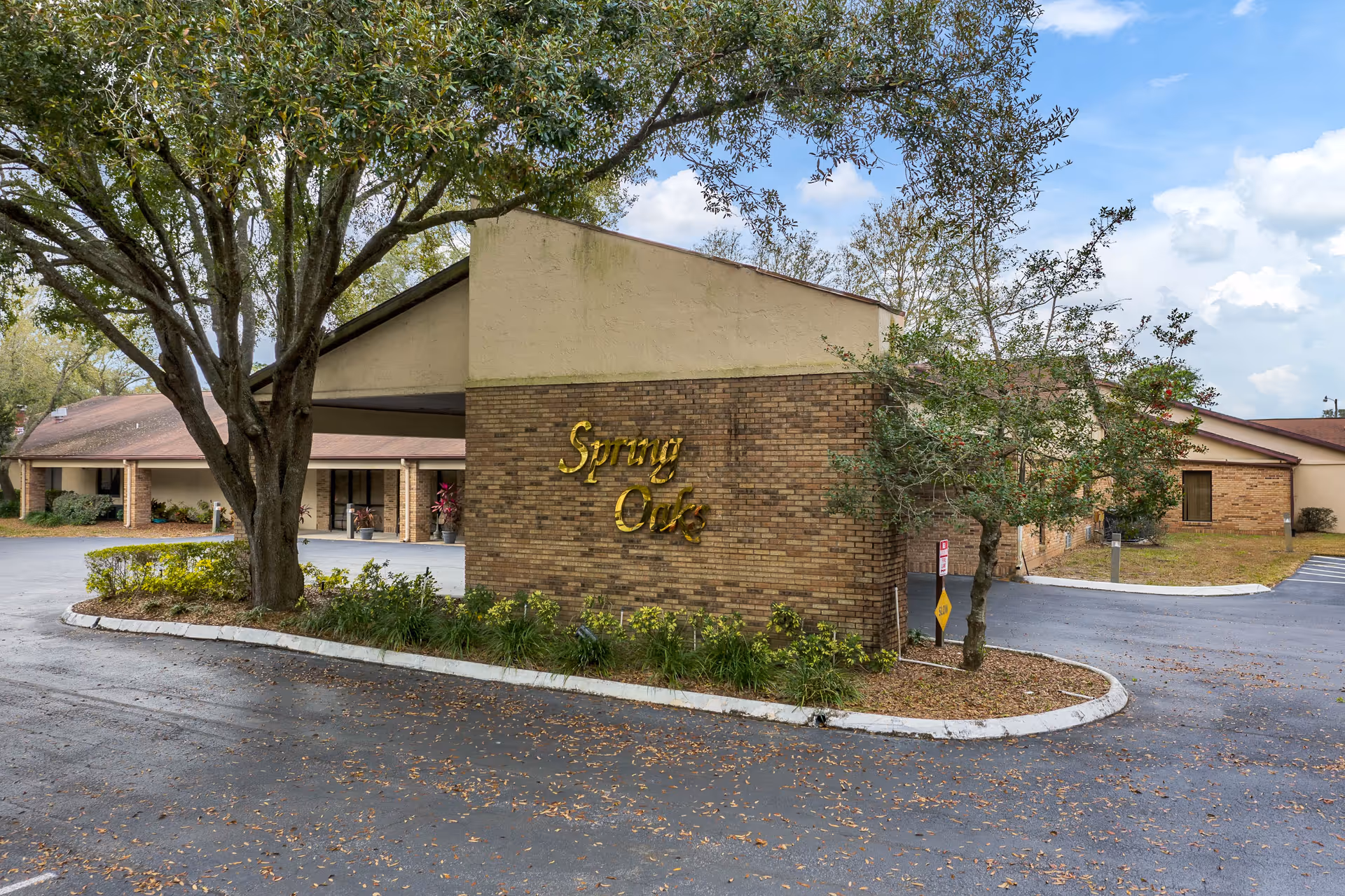 Exterior view of the Spring Oaks facility showing a brick building with a covered entrance, surrounded by trees and a paved driveway.