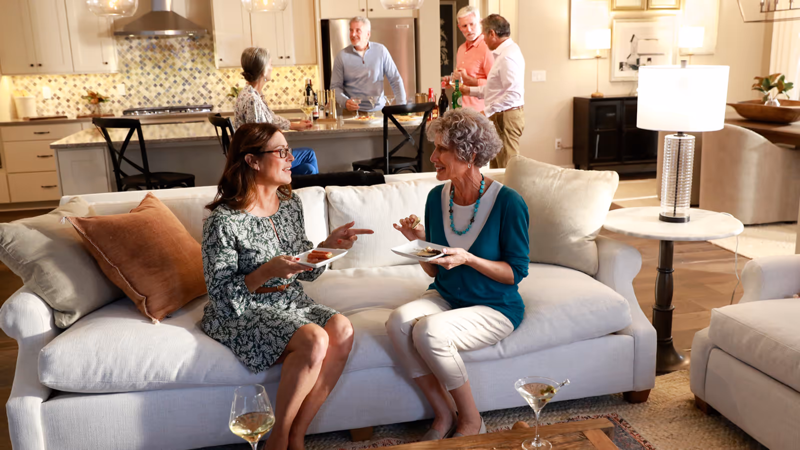 Two women sitting on a white couch in a living room, enjoying food and drinks while conversing. In the background, three people stand near a kitchen island with drinks, engaged in conversation. The room is warmly lit with modern decor, including a lamp on a side table and a kitchen with white cabinets and a tiled backsplash.