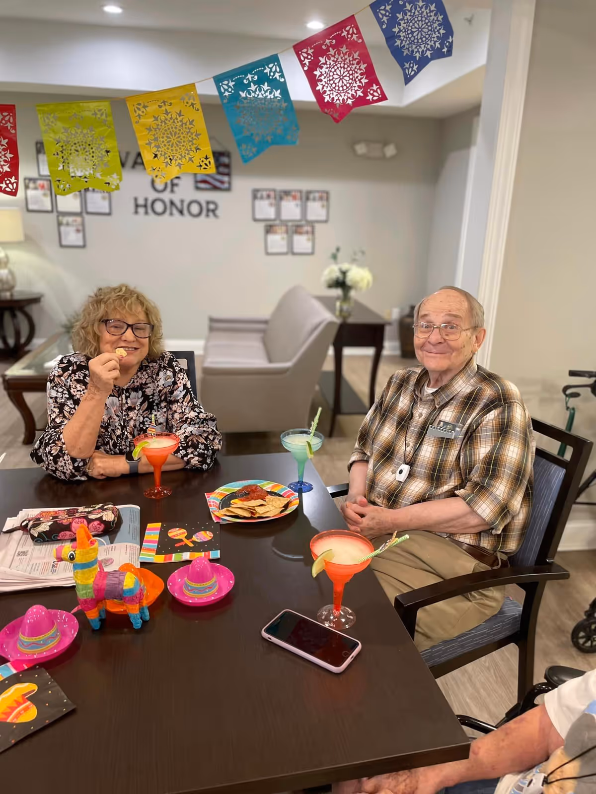 Two elderly people sitting at a table in a decorated room with colorful paper banners hanging above. The woman on the left is smiling and holding a chip, while the man on the right is smiling with his hands folded. On the table are festive decorations including small sombreros, a colorful piñata, plates with chips and salsa, and three margarita glasses with straws and lime wedges. The background shows a beige couch, a side table with flowers, and a wall with plaques and the words 'WALL OF HONOR'.