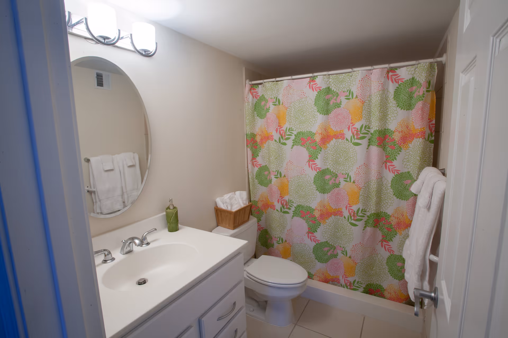 A small bathroom with a white vanity and sink, an oval mirror above the sink, a light fixture with two lights, a toilet next to the vanity, and a shower with a colorful floral shower curtain. White towels are hanging on a towel rack and a basket with rolled towels is placed on the toilet tank.