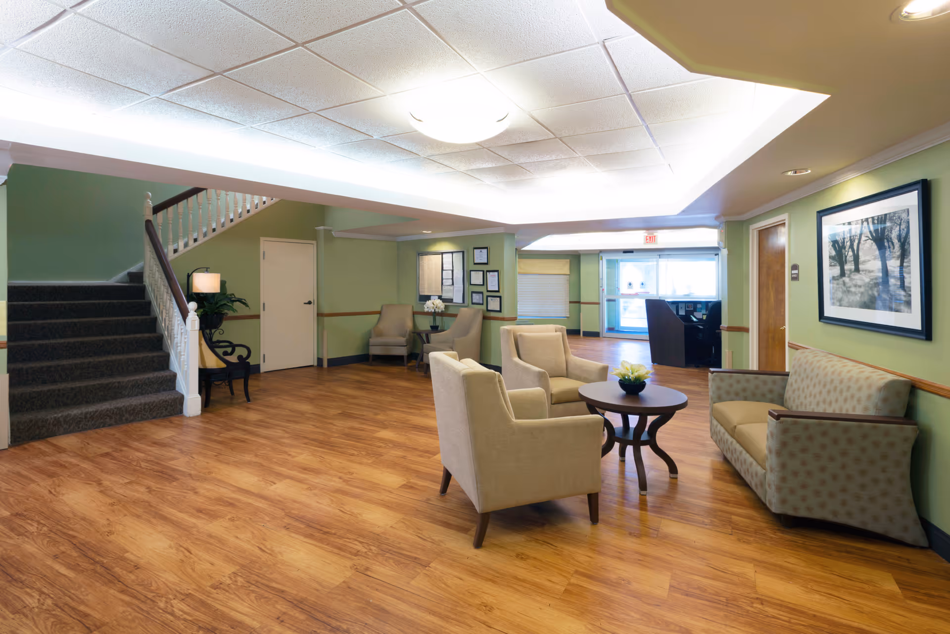 A spacious and well-lit common area with wooden flooring, light green walls, and a white ceiling with recessed lighting. The room features a seating arrangement with two beige armchairs and a patterned sofa around a round wooden table with a small plant centerpiece. There is a staircase with a dark carpet and white railing on the left side, a small table with a lamp and plant near the stairs, and a reception desk visible in the background near the entrance door.