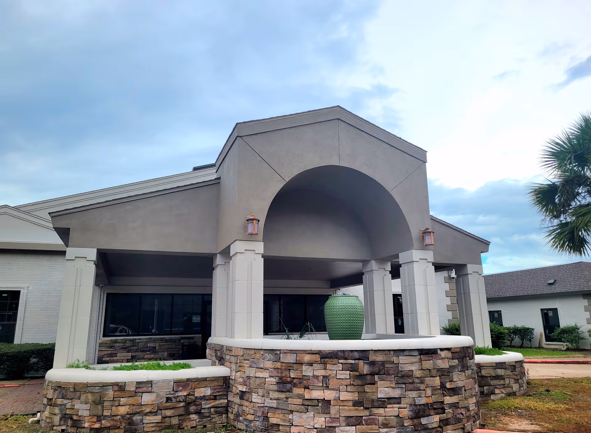 Exterior view of a building entrance with a covered porch supported by columns. The front features a stone wall with a large green decorative vase in the center. The sky is cloudy and there are some palm trees and shrubs around the building.