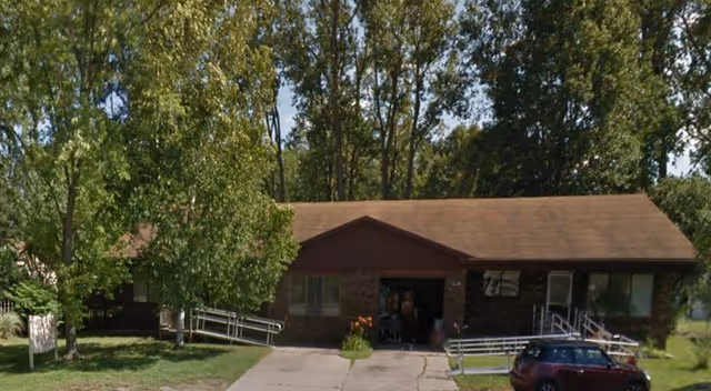 Single-story brick building with a brown roof surrounded by trees. There are two wheelchair ramps leading to the entrances on either side of the building. A small car is parked in front on the right side. The area is grassy with a concrete driveway in the center.