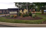 Outdoor view of the entrance area to Manor At St Luke Village, featuring a landscaped garden bed with flowers and plants surrounding a sign displaying the facility's name. A large wooden cross and a bench are visible in the background near the building.