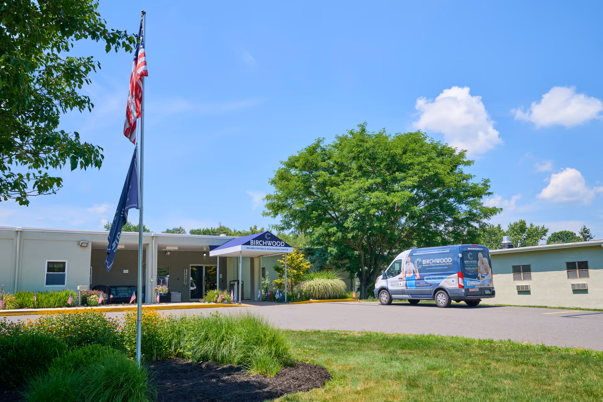 Exterior view of Birchwood Rehabilitation and Healthcare Center on a sunny day with a blue sky and scattered clouds. The building is single-story with a covered entrance displaying the facility's name. An American flag and another flag are flying on a flagpole in front of the building. A van with Birchwood branding is parked near the entrance. There are green trees, shrubs, and a well-maintained lawn surrounding the area.