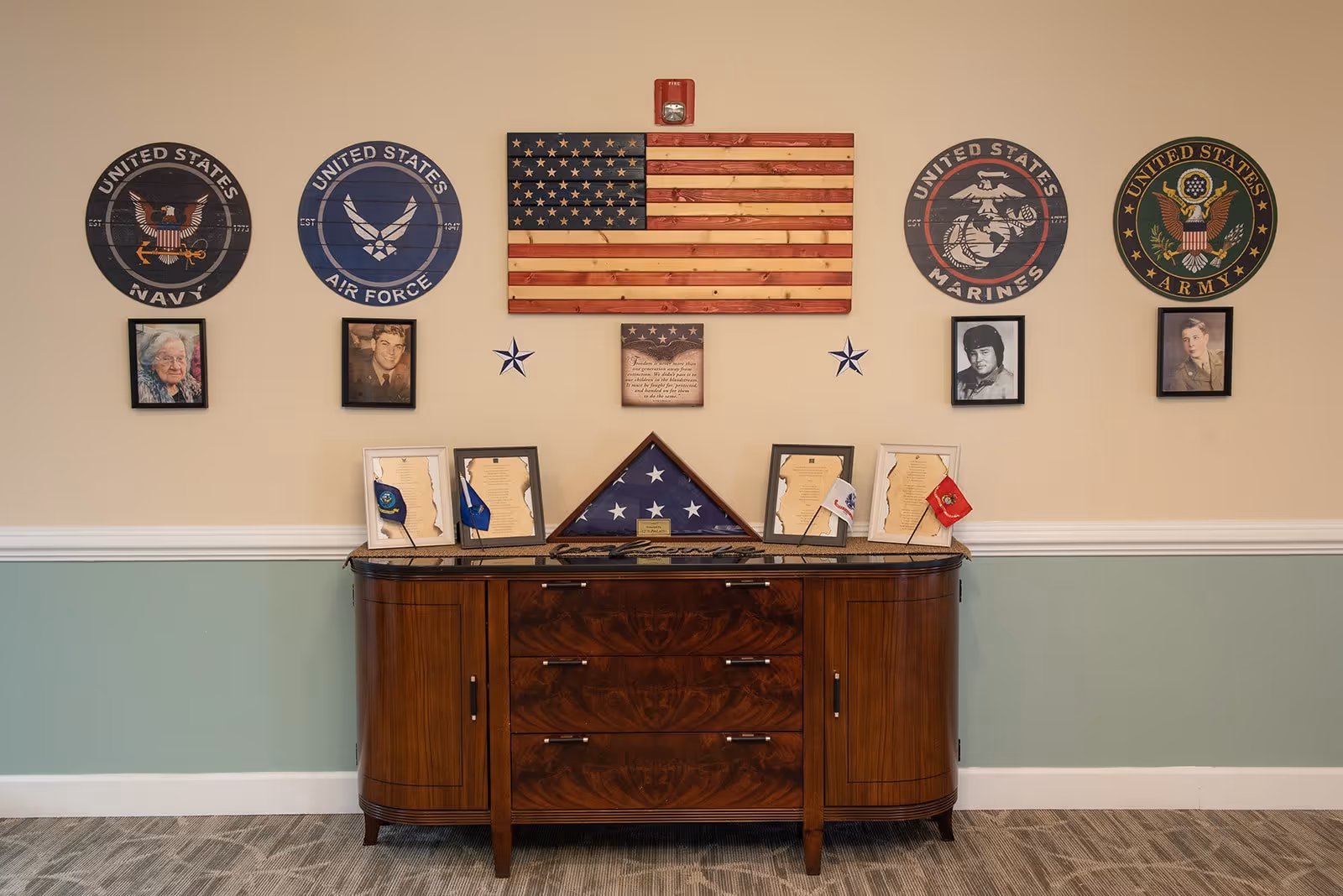 A memorial display in a senior living facility featuring a wooden American flag mounted on a beige wall above a wooden cabinet. On the wall are four circular plaques representing the United States Navy, Air Force, Marines, and Army, each accompanied by a framed photograph of a veteran. The cabinet holds framed documents, small flags, and a folded American flag in a triangular display case. The lower half of the wall is painted green with white trim, and the floor is carpeted.
