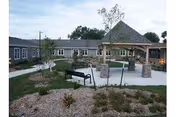 Outdoor courtyard area of a senior living facility with a gazebo, benches, paved walkways, and landscaped garden beds with small plants and mulch.